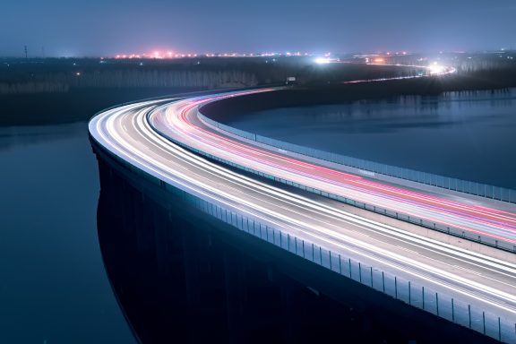 Photo of bridge at night with light trails from cars on it with city in the distance
