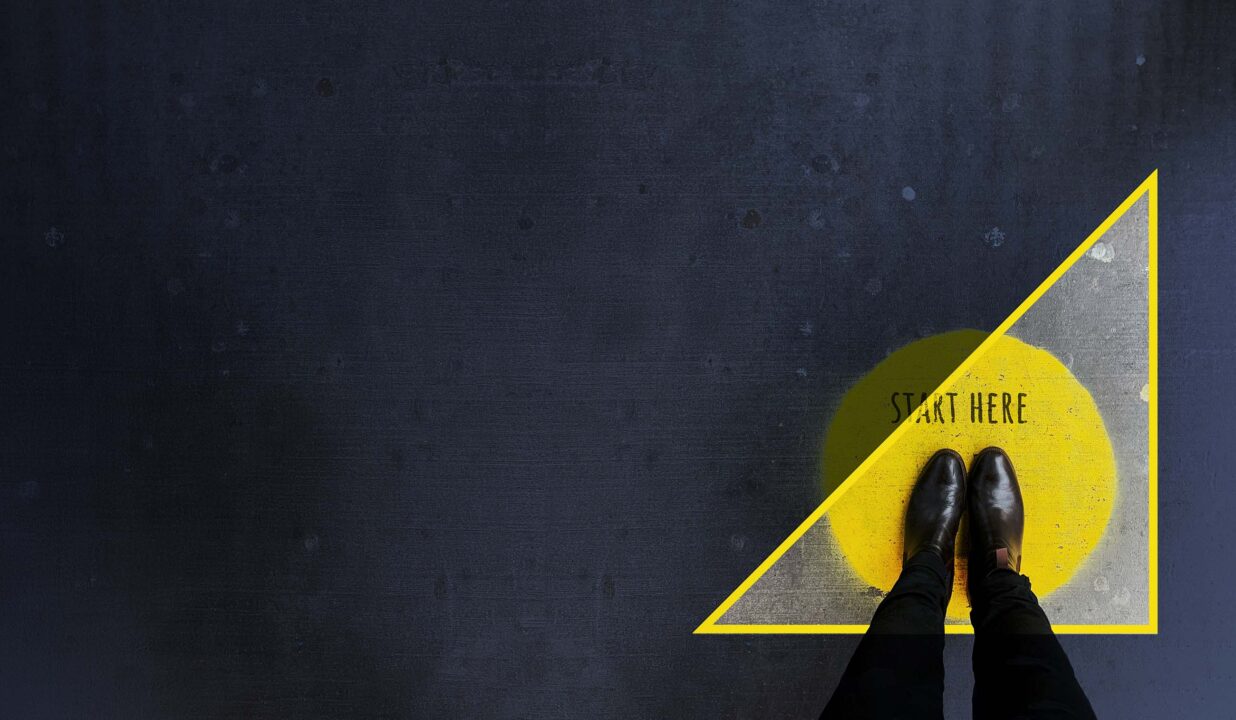 A downward facing shot of a businessman's slacks and shoes. They stand in a yellow painted circle on the concrete, with the words "Start Here" stenciled in black.